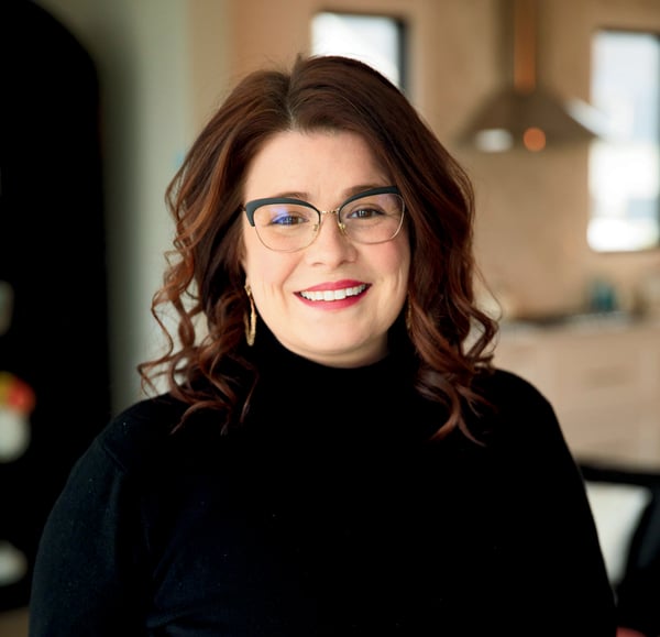 Portrait of Vikki Duke, a smiling woman with shoulder-length wavy brown hair, wearing glasses and a black turtleneck. She stands in a warmly lit modern kitchen. Vikki is the founder of Insomnia Mom, a Realtor, entrepreneur, and time management expert. She helps busy parents and professionals outsource tasks and reclaim control of their schedules using her Elevate & Delegate model.